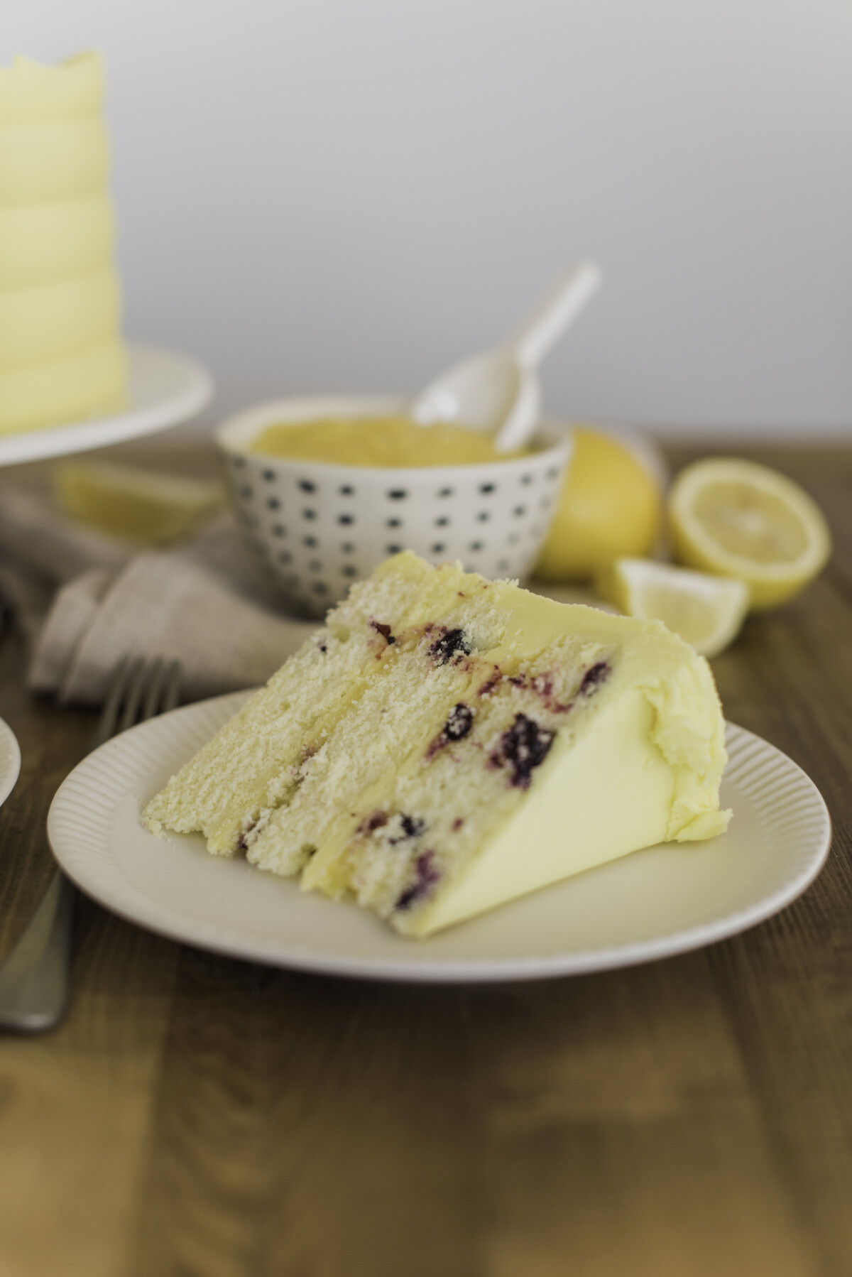 A slice of lemon blueberry cake on a plate with lemon curd and lemons nearby.