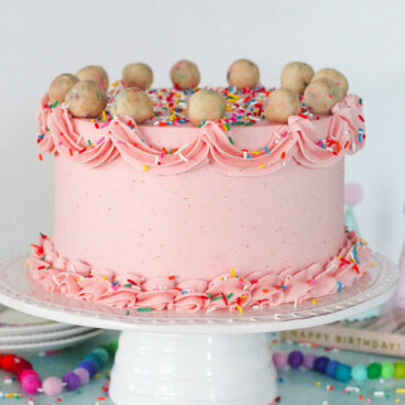 Pink cake on a cake stand with piping designs on the outside and cookie dough balls on top.