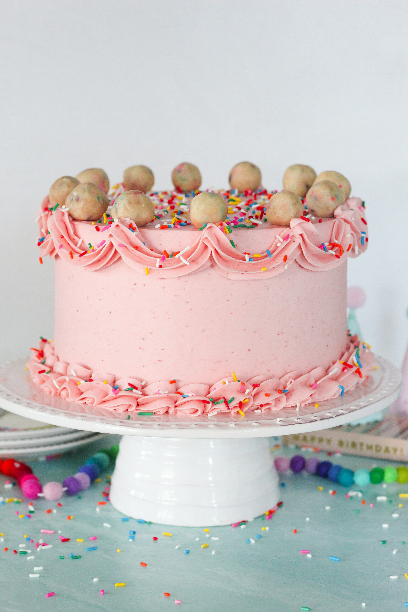 Pink cake on a cake stand with piping designs on the outside and cookie dough balls on top.